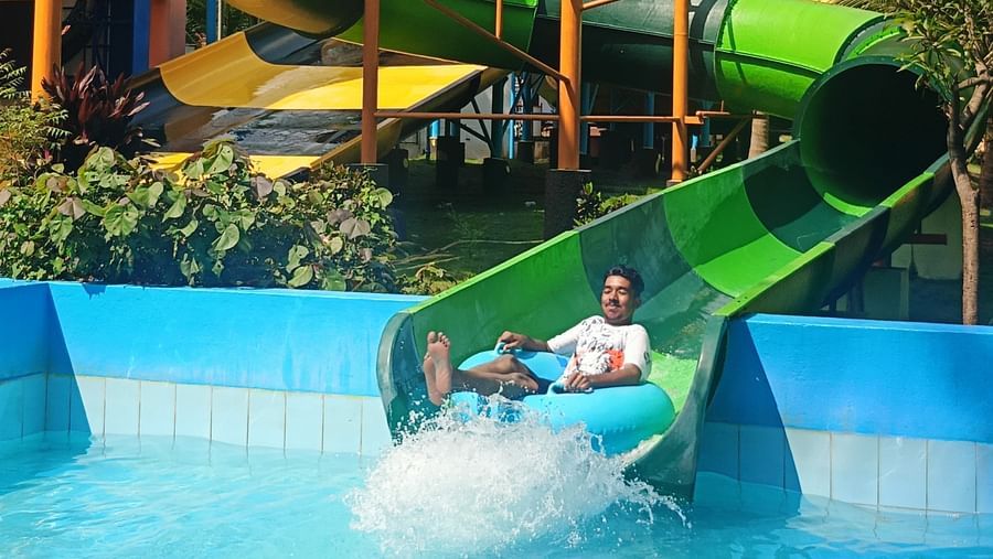 A rider splashes into the pool at the end of a green water slide, surrounded by palm trees and mountains.