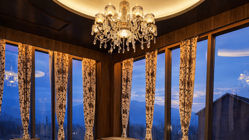 A dining space with circular seating arrangement at Mountnoor Sarovar Premiere. The space includes a curved sofa, armchairs, and a chandelier, all set against tall windows overlooking the valley at dusk.