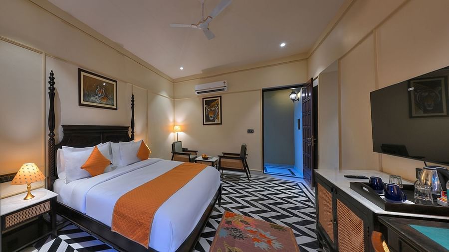An interior view of Superior Cottage featuring a dark wood four-poster bed with white linens and orange accents, a flat-screen TV, a desk, and artwork depicting tigers on the walls - Singhvilas, Ranthambore