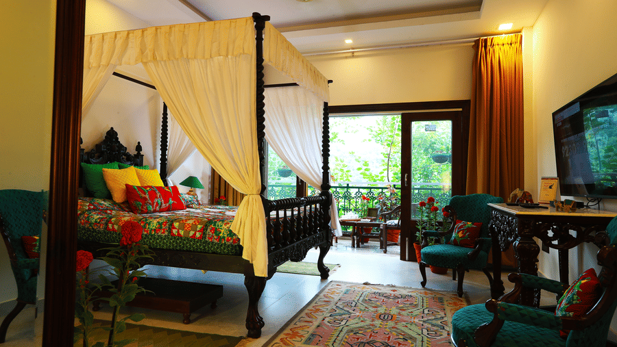 The Parakeet Room at Shaheen Bagh featuring a bed with canopy, wooden tables and chairs with plush cushions, and a balcony facing the lush greenery of the property.
