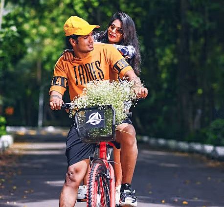A man riding a bicycle and a woman sitting behind him, looking at each other with flowers in the cycle basket, surrounded by greenary