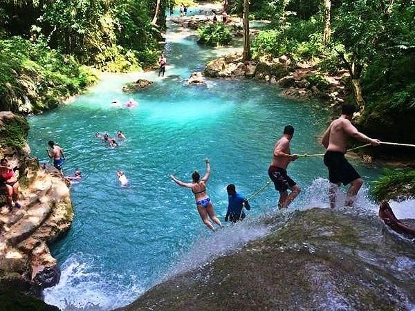 People swing from ropes and swim in a beautiful turquoise river surrounded by lush green jungle, enjoying a waterfall.