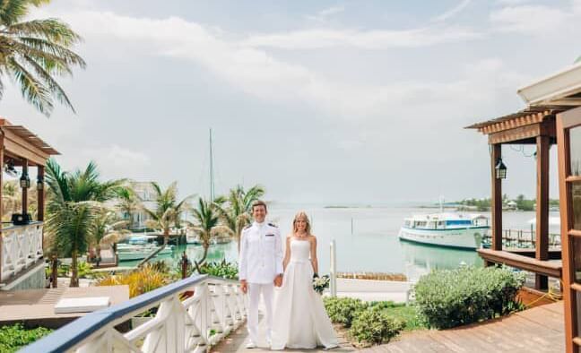 Two people in wedding attire walk on a deck lined with palm trees near boats and water at Abaco Inn.