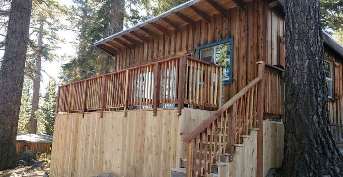 An exterior of a rustic, wooden cabin at the Kit Carson Lodge, featuring a staircase leading to a balcony and surrounded by trees.