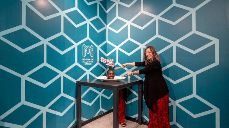 A woman stands next to a table where a person appears to have their head on a plate as part of a visual trick against a wall with geometric patterns.