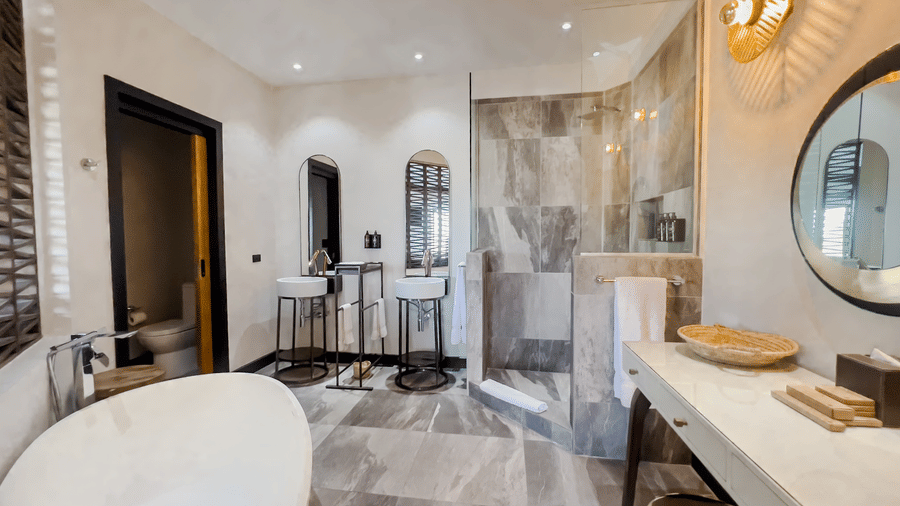 A bathroom at Tamarind Hills Resort and Villas, Antigua featuring a freestanding oval bathtub, a glass-enclosed shower, double sinks with plumbing fixtures, and a lighting set up and marble-style flooring.
