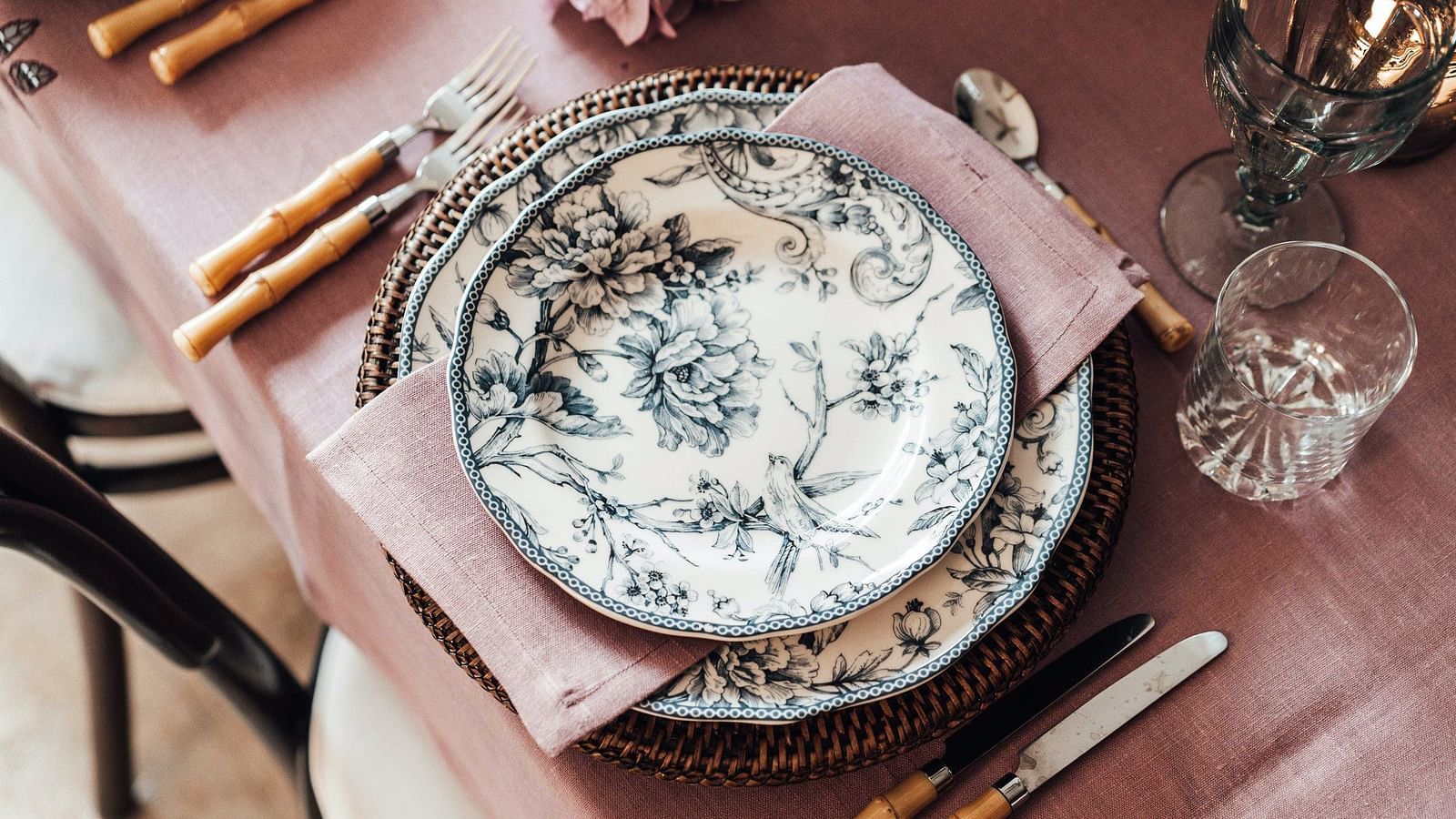 Detail of a dining place setting with a bamboo-handled fork and knife, a floral-patterned plate, on a raffia place mat and rose-coloured linen
