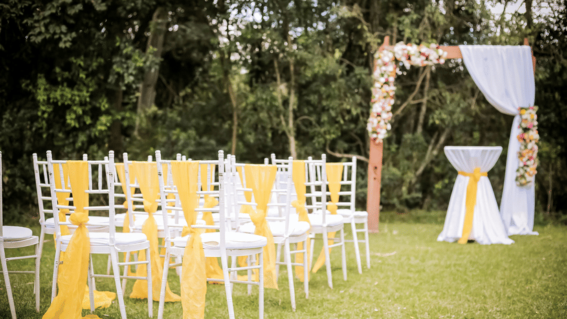 An outdoor event setting with rows of white chairs with yellow sashes, facing a decorated archway.