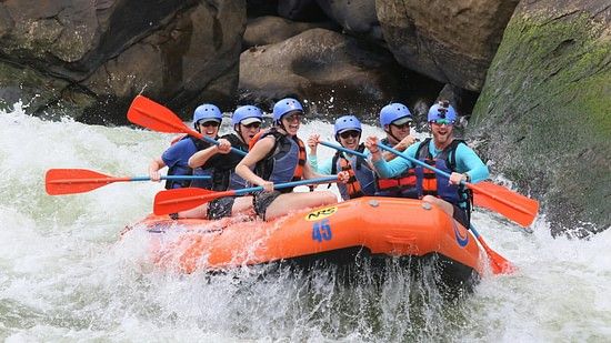 Another group of people in blue life jackets and helmets navigating through white water rapids in an orange raft, with large rocks and turbulent water around.