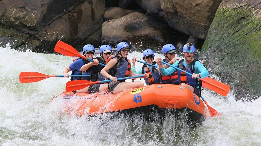 Another group of people in blue life jackets and helmets navigating through white water rapids in an orange raft, with large rocks and turbulent water around.