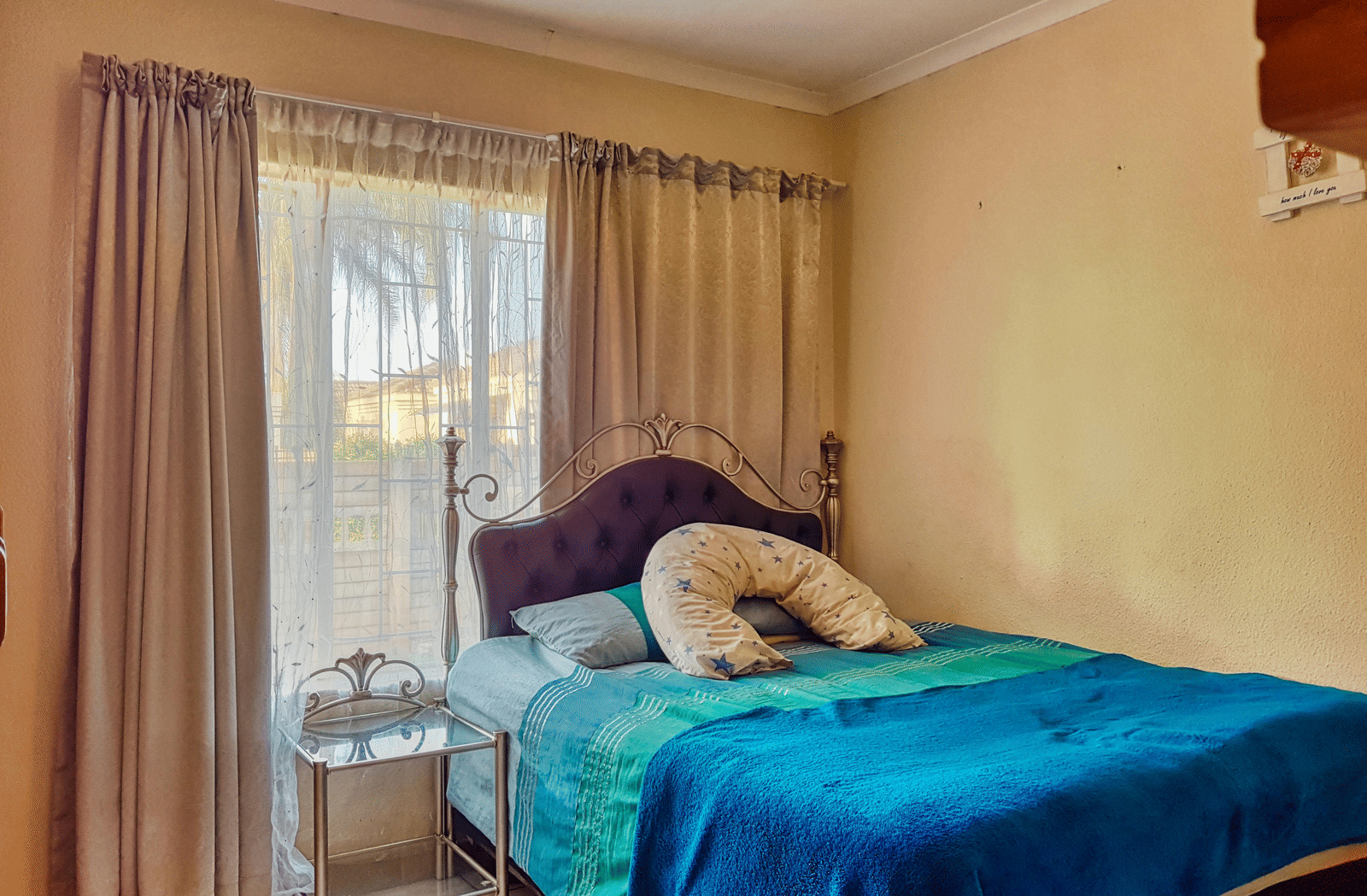 A bedroom featuring a double bed with blue sheets spread neatly over it with beige curtains covering the window behind it.