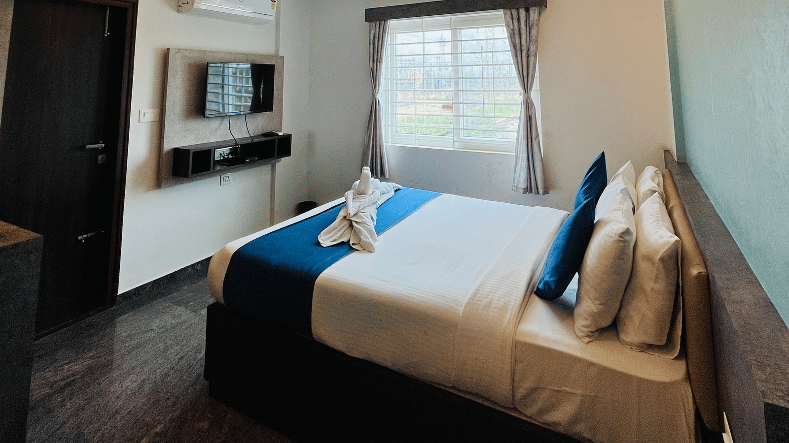 Queen bed under a recessed ceiling with warm cove lighting and a wall mounted TV in the Standard Balcony Room at Value Inn, Bangalore Airport