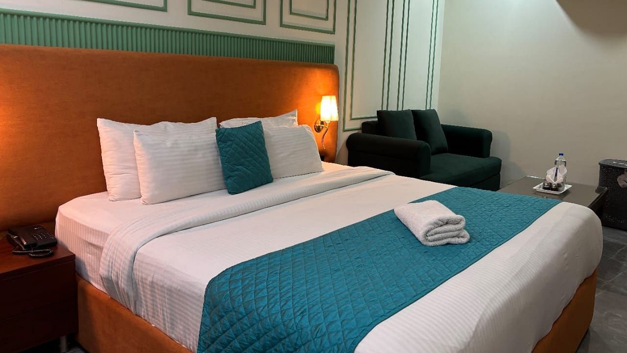 A well-lit room with a double bed dressed in white and blue, and a dark armchair - Asian Suites IFFCO Metro, Gurgaon