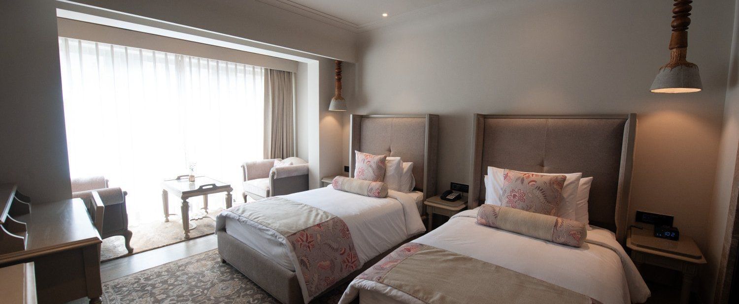 A wide-angle shot of a Premium Twin Room at The Evren, Vagator, showing two comfortable single beds, a desk, and a seating area with two armchairs and a coffee table near the large window.