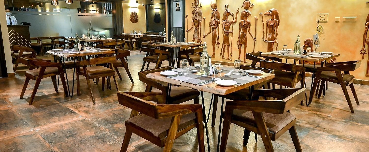 Another angle of the restaurant dining space, focusing on a few tables set with placemats and cutlery, against a backdrop of unique wall art - Sea Hills Hotels, Sri Vijaya Puram (Port Blair)