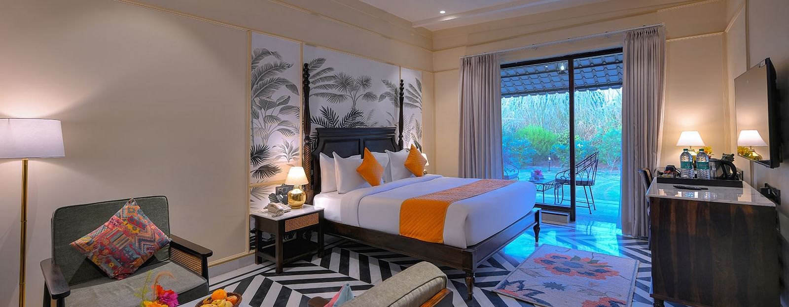 Superior Cottage featuring a four-poster bed with white linens, a colourful patterned runner and pillows, a patterned armchair, a wall-mounted TV, and sliding glass doors leading to a balcony - Singhvilas, Ranthambore