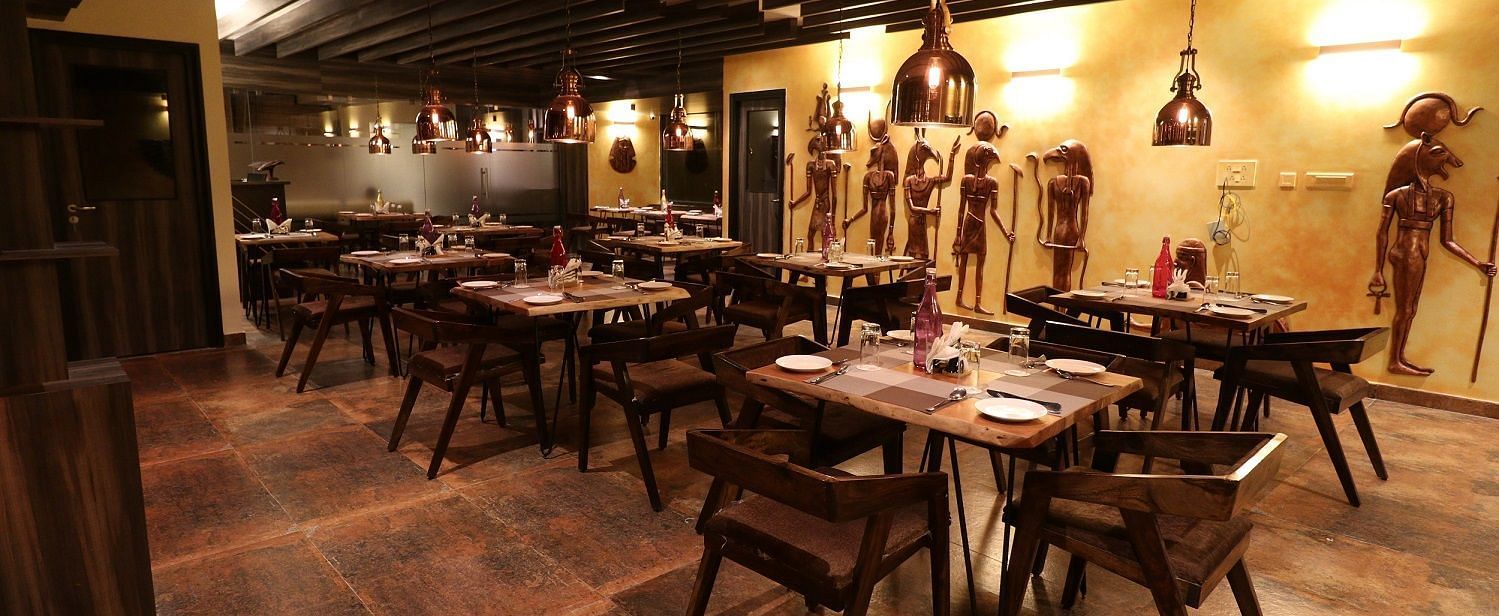 A central perspective of a restaurant dining area, showing multiple set tables with wooden chairs, warm lighting from hanging fixtures, and distinct wall decorations - Sea Hills Hotels, Sri Vijaya Puram (Port Blair)