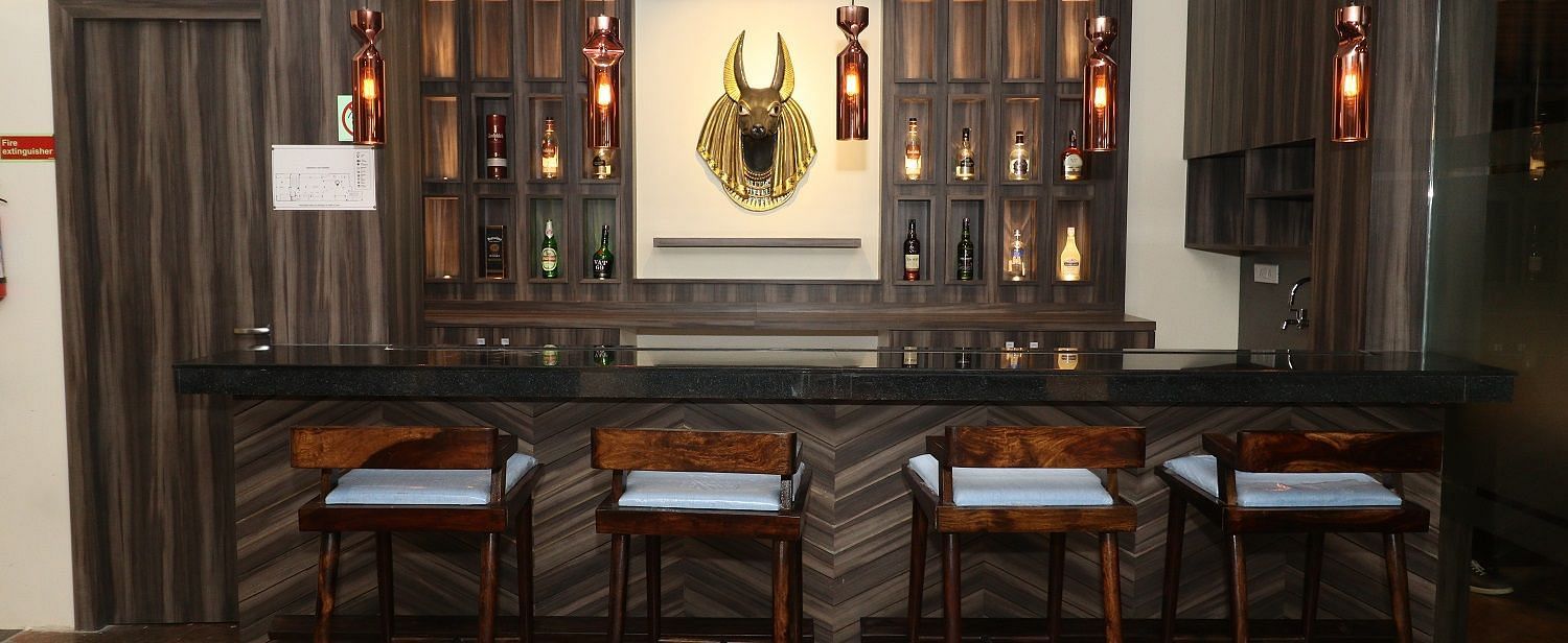 A well-stocked bar area with dark wooden counters, high bar stools, and bottles displayed on shelves, illuminated by hanging lights - Sea Hills Hotels, Sri Vijaya Puram (Port Blair)