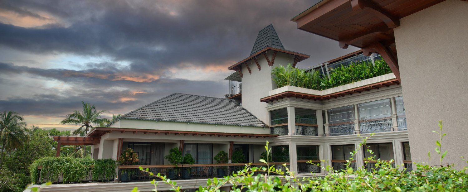 An architectural shot of the exterior of The Evren, Vagator, showing the elegant facade, balconies, and the lush green surroundings. The image is taken from a vantage point that captures the building's design against a dramatic sky.