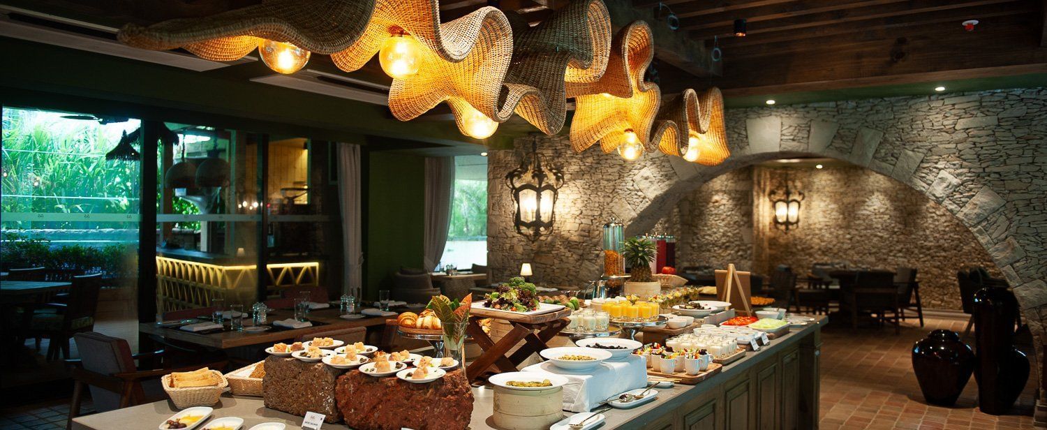 A lavish breakfast buffet spread at Mila Restaurant at The Evren, Vagator, featuring a variety of dishes on a large central island. The restaurant has a rustic yet modern ambiance with woven light fixtures, a stone archway, and a view of the outdoors.