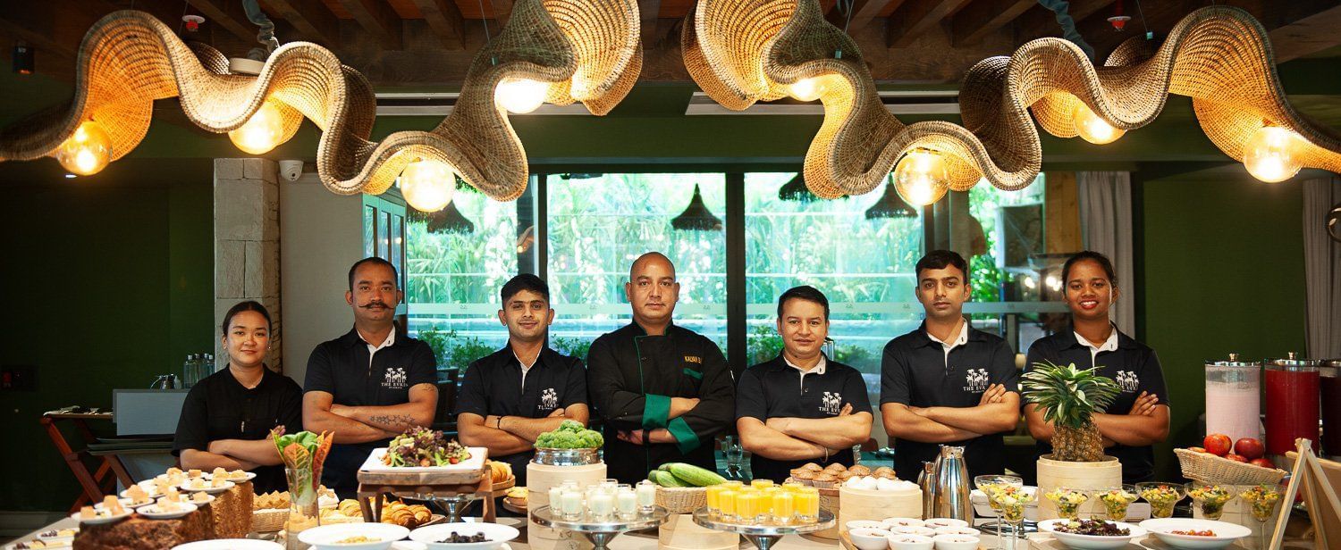  The culinary and service team of Mila Restaurant at The Evren, Vagator, standing proudly behind a beautifully presented buffet spread. The team is smiling and wearing their uniforms, with the head chef in the center.