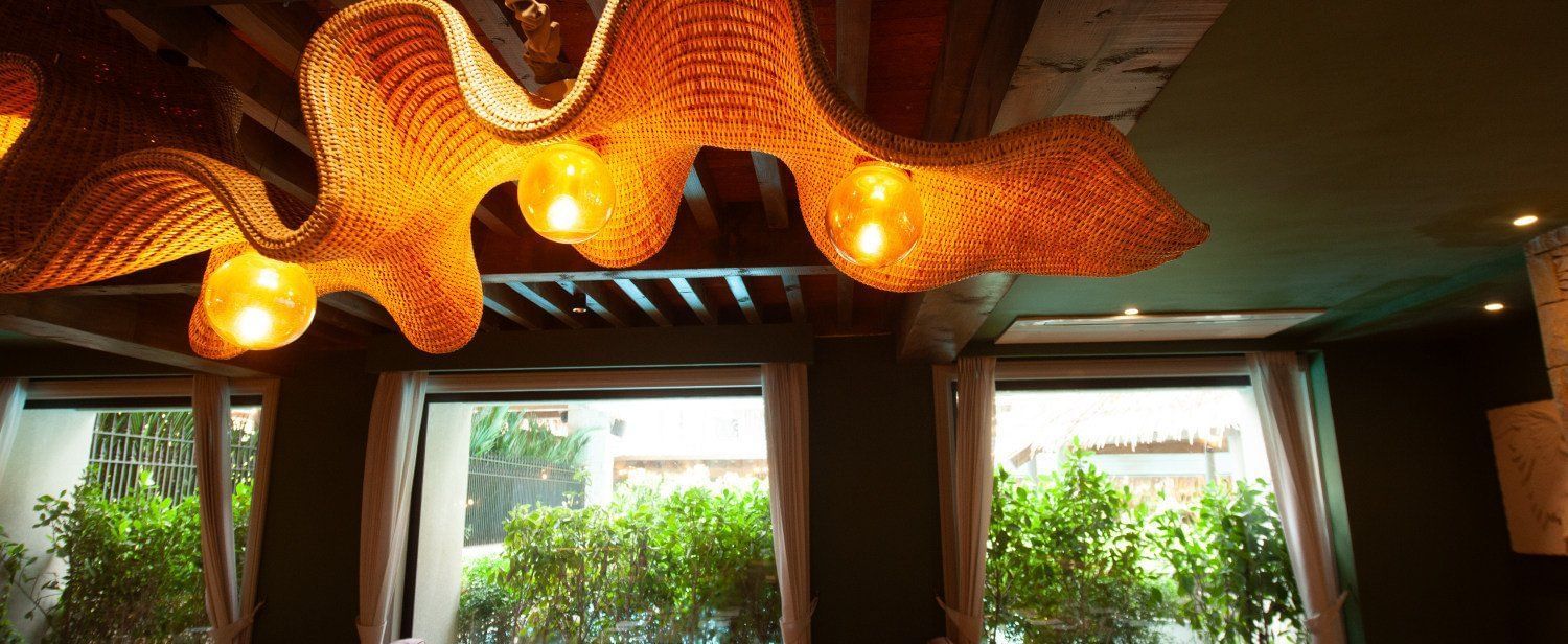 A detailed shot of the interior of Mila Restaurant at The Evren, Vagator, highlighting the unique, woven light fixtures hanging from the wooden ceiling. The restaurant has large windows with a view of greenery and comfortable seating.