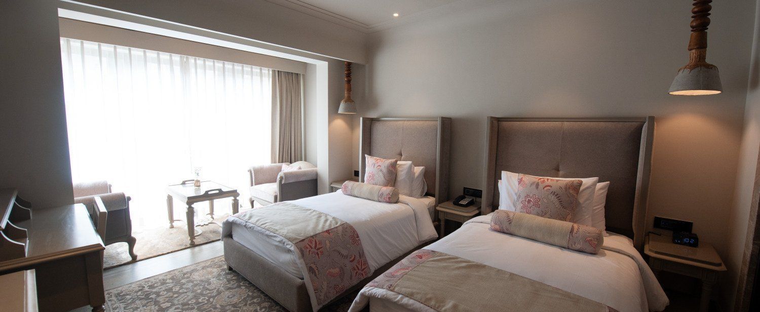 A wide-angle shot of a Premium Twin Room at The Evren, Vagator, showing two comfortable single beds, a desk, and a seating area with two armchairs and a coffee table near the large window.