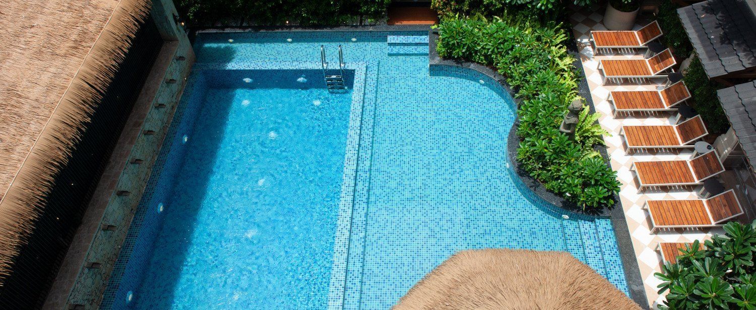 An aerial view of the outdoor swimming pool at The Evren, Vagator, featuring bright blue mosaic tiles, a built-in seating area, and a row of lounge chairs shaded by a large straw umbrella. The pool is surrounded by lush green plants and is overlooked by th