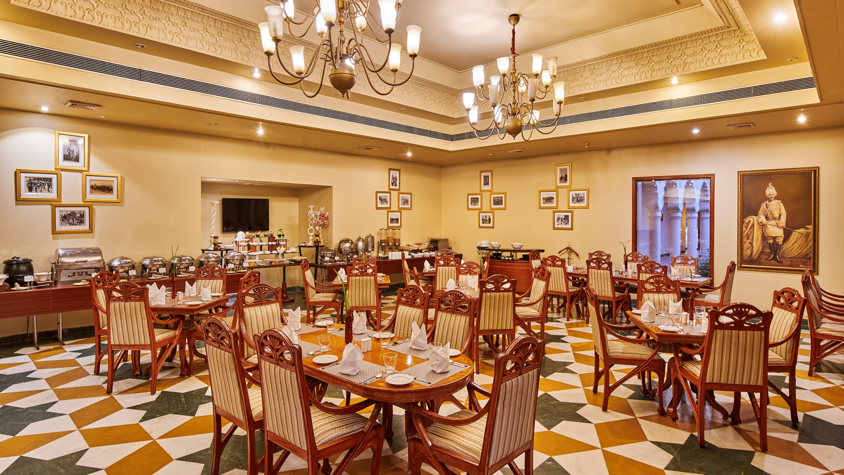 Sir P restaurant at The Ummed Jodhpur, Palace Resort and Spa, with patterned tiled flooring, neatly set round tables, upholstered chairs, chandeliers and a buffet counter along the wall.