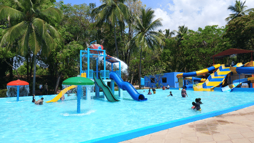 A water park scene with colourful slides, water play structures, and umbrellas, surrounded by lush palm trees and greenery, with children and families enjoying the pool under a clear blue sky.