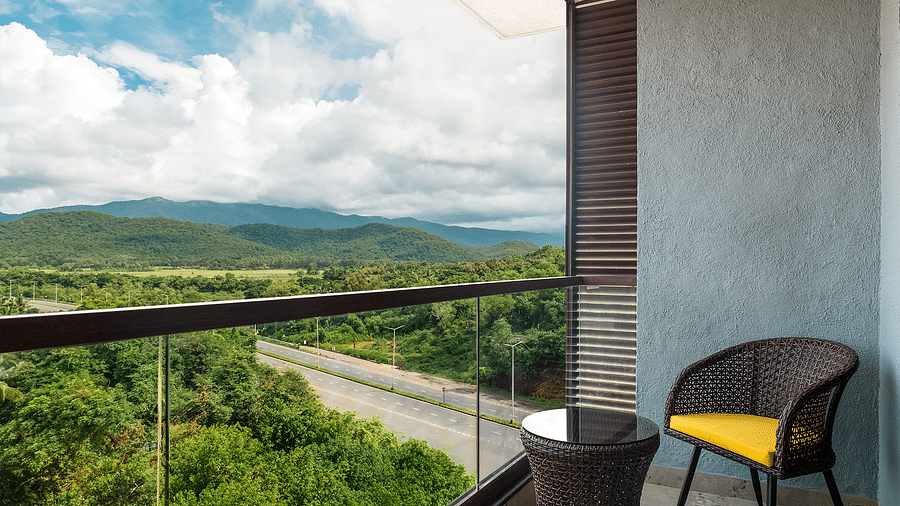  A chair and coffee table on the balcony of one of the rooms offer a view of lush trees and mountains under a cloudy sky at Cloud9 Sarovar Premiere, Palolem Goa