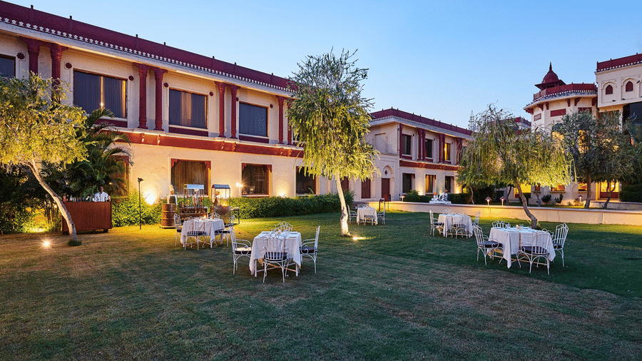 Evening view of the expansive lawn at The Ummed Jodhpur, Palace Resort and Spa, with scattered white outdoor dining setups, manicured grass, trees and the softly lit palatial façade under a clear sky.