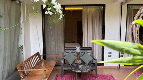 An outdoor terrace features a wooden swing, 2 chairs, a coffee table, and hanging white bougainvillea flowers by a glass door at Bougainvillea, Alibaug.