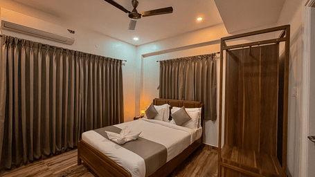 Wide veiw of a bedroom in the Coral 4BHK of Kove Smart Stays featuring wooden flooring, plush bedding, a wardrobe unit, and closed curtains above the headboard of the bed, and one whole wall of the room.