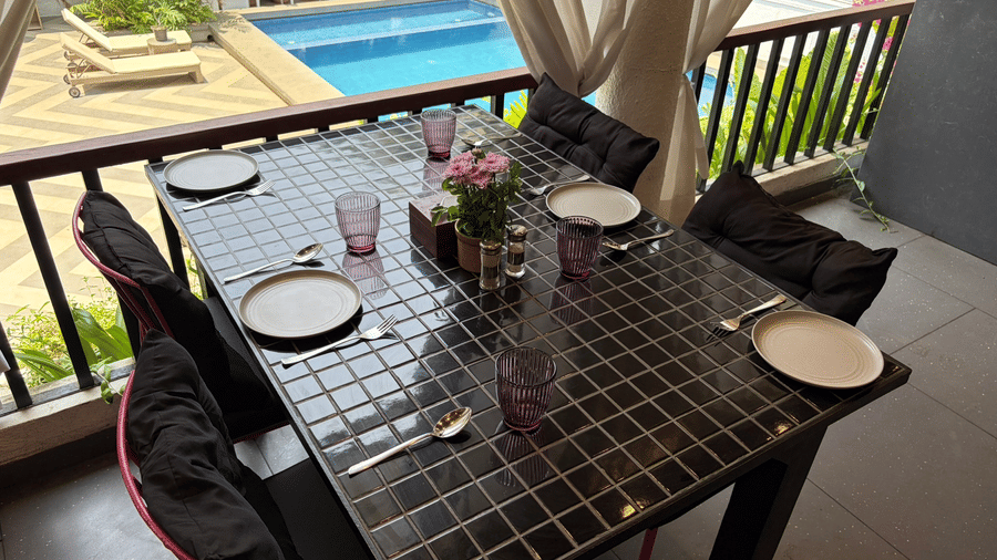 A tiled dining table is set with plates, glasses, and cushions on a balcony overlooking a swimming pool at Bougainvillea, Alibaug.