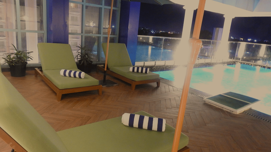 A rooftop swimming pool area at Sands Point Hotel, Pondicherry, featuring green sun loungers with rolled striped towels, large green patio umbrellas, and a wooden deck.