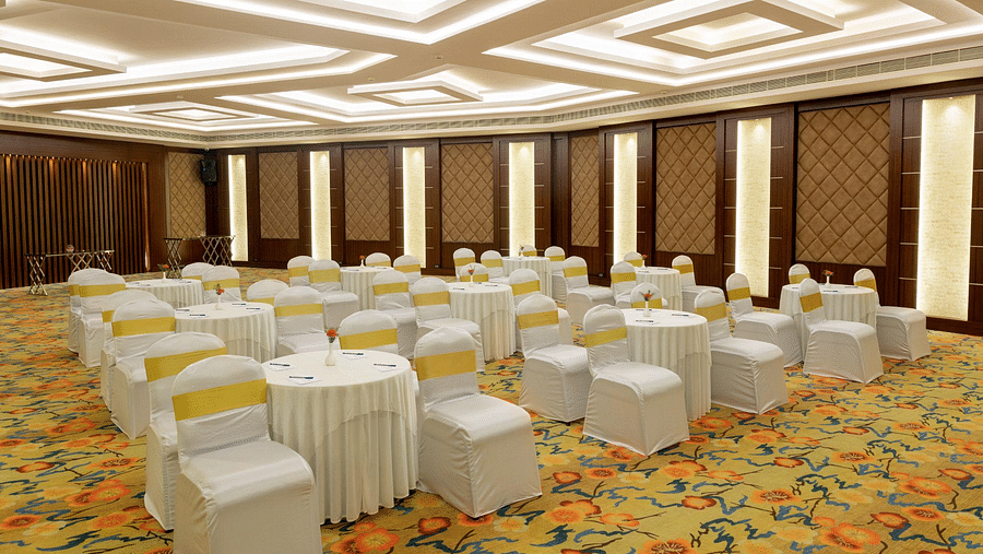 Banquet hall with round tables draped in white, chairs with yellow sashes, patterned carpet, and a geometrically designed ceiling - Jenneys Residency, Coimbatore