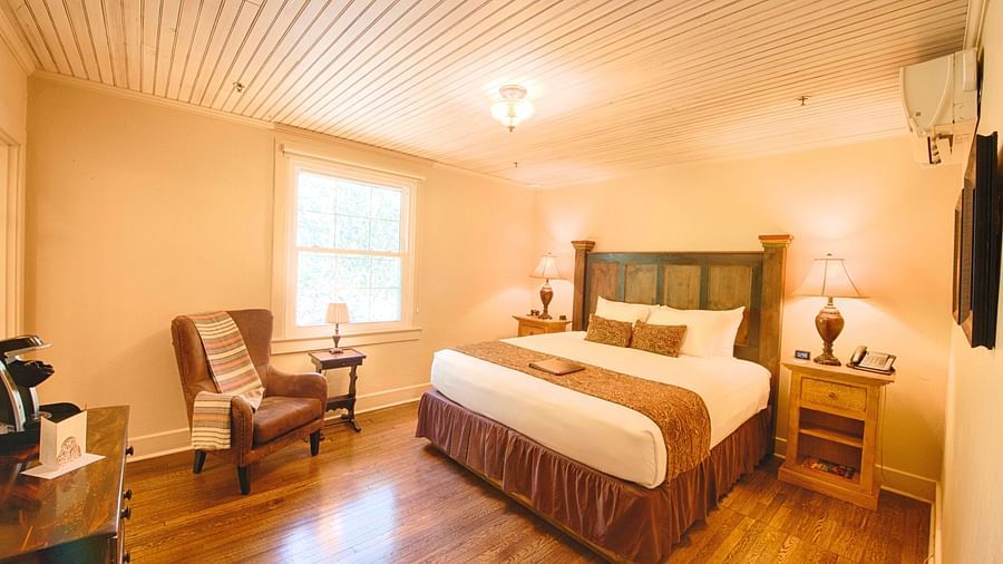 full view image of a brightly lit bedroom with a comfortable double bed with a heady headboard along with an armchair - Historic Tapoco Lodge