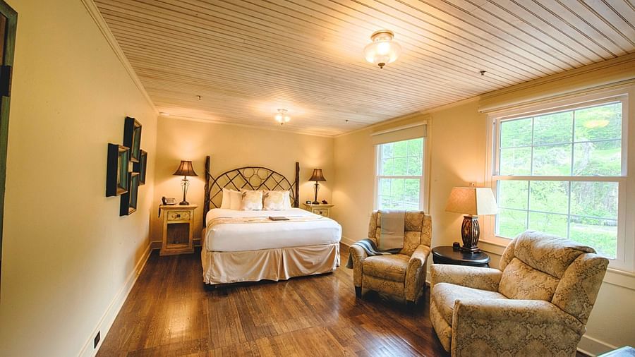 full view image of a room with wide windows featuring a cozy double bed and comfortable cushioned armchairs in front of it - Historic Tapoco Lodge