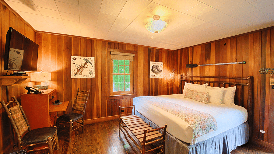a room with wooden walls featuring a double bed with soft pillows arranged over it along with a wall mounted television right in front of it - Historic Tapoco Lodge