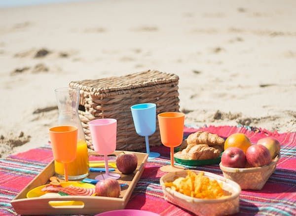 A beach picnic setup with food and drinks laid out on a checkered blanket, with the ocean in the background.