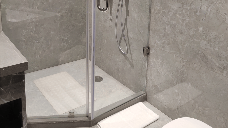 A bathroom at Anant Mahal, Jaipur featuring a glass shower enclosure, grey tiled walls, and white towels on a metal rack.