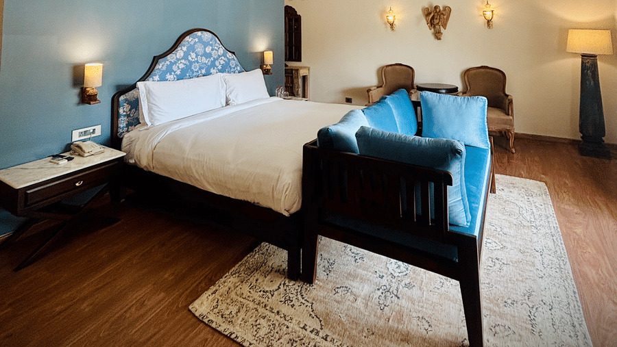 Vana Junior Suite at Surpura Bagh featuring a double size bed, bedside table with lamp on either side, two sofa chairs with centre table, a tall lamp, and a long sofa attached to the bed.