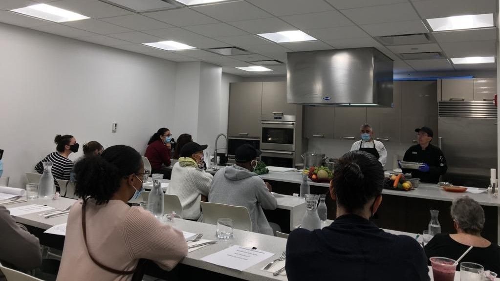 A group of people attending a cooking class, watching chefs demonstrate techniques at the front of a modern kitchen classroom at YO1 Longevity & Health Resorts, Catskills