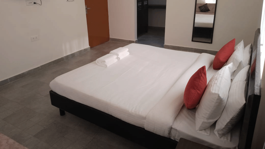 Bright guest room featuring a king size bed, red cushions, wall mounted television and dressing mirror.