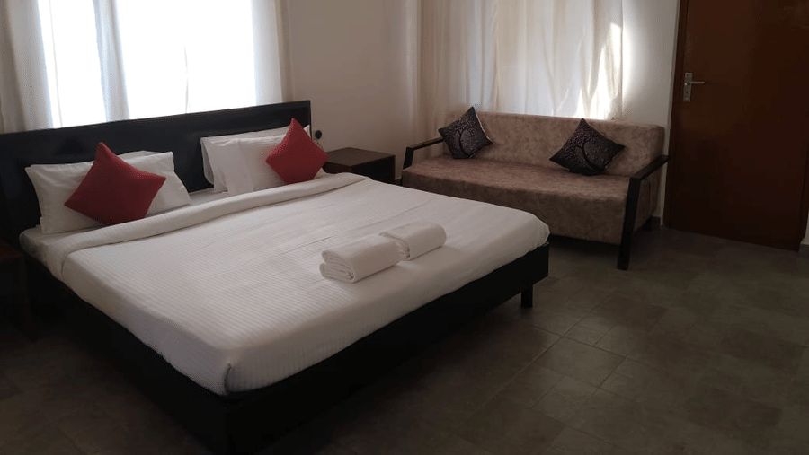 Guest room with a king size bed, red accent cushions, bedside seating and large curtained windows.