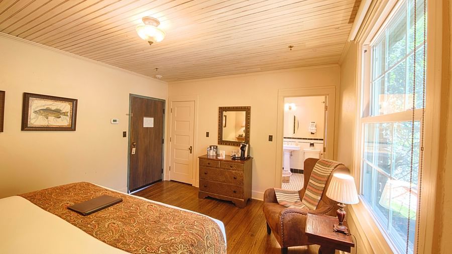 a cozy bedroom with a cushioned rustic armchair along with a small side table with a table lamp and a double bed in the room - Historic Tapoco Lodge