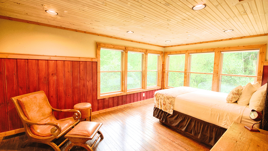 corner view image of a vibrant bedroom with an armchair and a bed overlooking the beautiful green forest - Historic Tapoco Lodge