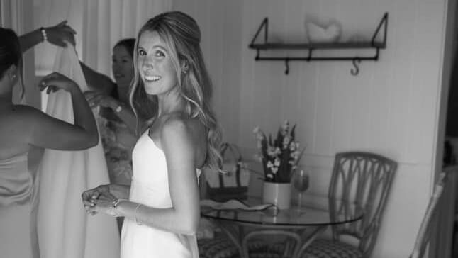 Person in a wedding dress stands indoors holding a bouquet, with another person adjusting the dress, chair and wall art in the background at Abaco Inn.