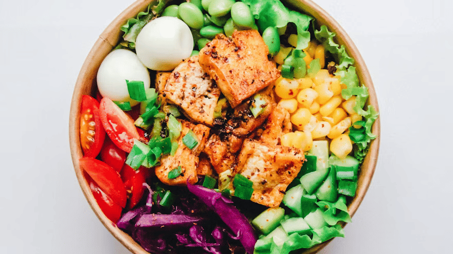 Overhead view of a salad bowl with spiced tofu cubes, boiled eggs, edamame, sweet corn, cucumber, cherry tomatoes, red cabbage, and lettuce.